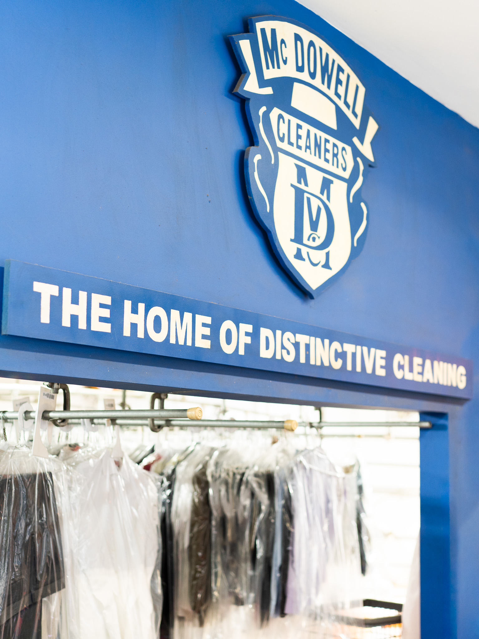 Blue sign above a clothing rack reads "McDowell Cleaners" and "The Home of Distinctive Cleaning"; dry-cleaned clothes hang below.