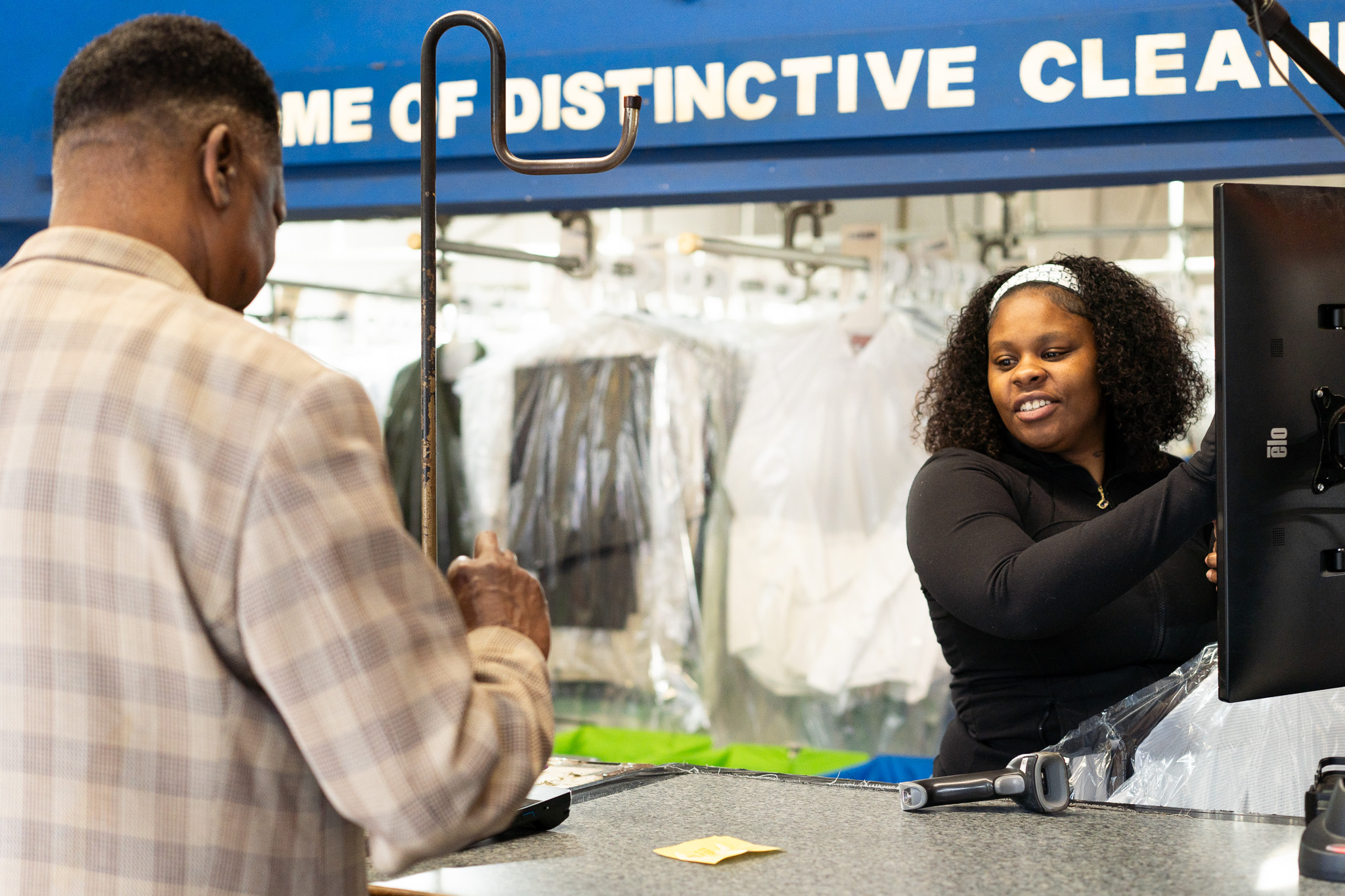 A woman smiles while assisting a customer at the counter of a dry cleaning business, with clothes hanging in plastic in the background.
