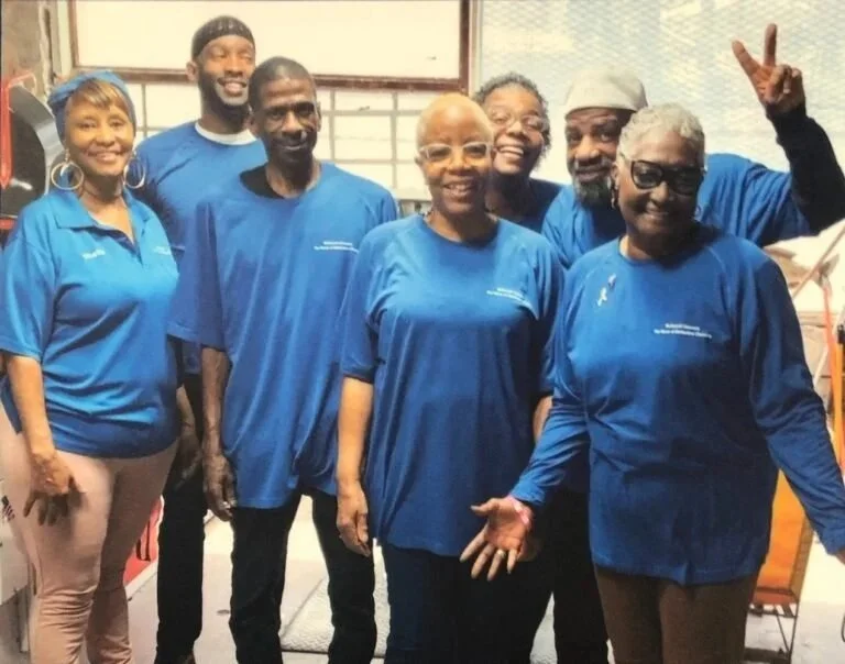 Seven adults stand indoors, smiling and wearing matching blue shirts, while one person in the back makes a peace sign.